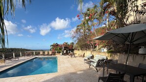 Área na beira da piscina com vista desobstruída para o oceano, cadeiras de descanso, uma mesa e um guarda-sol.