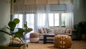 Uma sala de estar com sofá, apoio para os pés e mesa de centro. Há cortinas, uma planta e uma janela com persianas.
