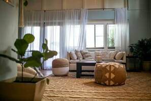 Ein Wohnzimmer mit einer Couch, einem Hocker und einem Couchtisch. Vorhanden sind Vorhänge, eine Pflanze und ein Fenster mit Jalousien.