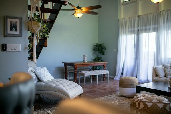 A cozy living room with a sofa, a coffee table, a staircase with macrame decor, a ceiling fan, and a dining area with a table and chairs. - Cruz Bay 1BR Loft, Pool, A/C, Backup Power + In-House Car Rental (Cruz Bay)