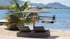 Een strandlounge met rieten stoelen en een geweven parasol, met uitzicht op een rustige zee en verre bergen.