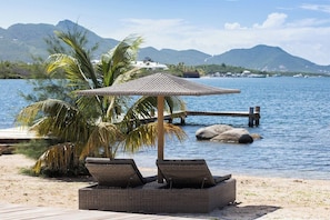 Eine Strandlounge mit Korbstühlen und einem gewobenen Sonnenschirm mit Blick auf ein ruhiges Meer und ferne Berge.
