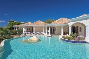 Un área junto a la piscina con una piscina cristalina, columnas blancas y sillones de descanso.