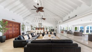 Ein großzügiger Wohnbereich mit einem großen, schwarzen Ledersofa, einem Couchtisch und Deckenventilatoren. Im Hintergrund befinden sich Kübelpflanzen und eine Holztür.
