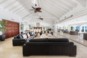 Ein großzügiger Wohnbereich mit einem großen, schwarzen Ledersofa, einem Couchtisch und Deckenventilatoren. Im Hintergrund befinden sich Kübelpflanzen und eine Holztür.