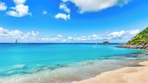 Ein Strand mit klarem türkisblauem Wasser, einem sandigen Ufer und einem Segelboot in der Ferne.