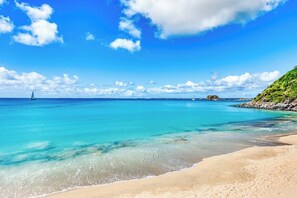 Ein Strand mit klarem türkisblauem Wasser, einem sandigen Ufer und einem Segelboot in der Ferne.
