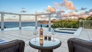Una terraza con una mesa de vidrio, dos sillas de mimbre, una botella y dos vasos. La terraza tiene vista a una piscina y un paisaje escénico del mar y las montañas.