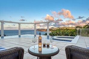 Eine Terrasse mit einem Glastisch, zwei Korbstühlen, einer Flasche und zwei Gläsern. Von der Terrasse hat man Blick auf einen Pool und eine malerische Aussicht auf das Meer und die Berge.