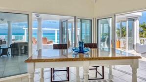 Amplia habitación con una gran mesa de vidrio y sillas, vista al océano y un balcón con mobiliario exterior.
