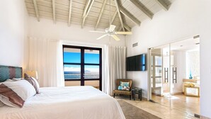 Een ruime slaapkamer met een groot bed, een plafondventilator, uitzicht op de oceaan door een schuifdeur en een aan de muur bevestigde televisie.