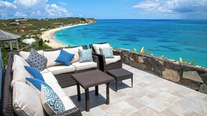 A patio with wicker furniture, a stone wall, and a view of a beach and ocean. - Libellule   Elevated Beachfront Villa (Saint Martin)