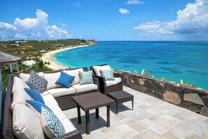 A patio with wicker furniture, a stone wall, and a view of a beach and ocean. - Libellule   Elevated Beachfront Villa (Saint Martin)