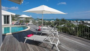 Eine Holzterrasse mit Pool, weißen Liegestühlen und Sonnenschirmen.