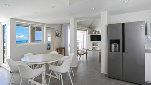 Eine moderne Küche mit einem Edelstahlkühlschrank, einem Essbereich mit weißen Stühlen und großen Fenstern mit Meerblick.