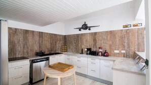 A modern kitchen with white cabinets, a wooden backsplash, a dishwasher, a coffee maker, and a wooden stool. - Mary's Beach Cottage: Seashore (Saint Martin)