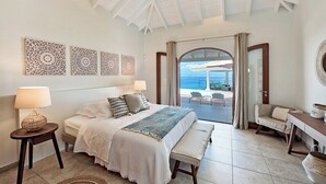 A bedroom with a bed, bedside tables, a bench, and a view of the ocean through a glass door. - Happy Bay   Classic French Creole Villa   Stunning Views (Saint Martin)