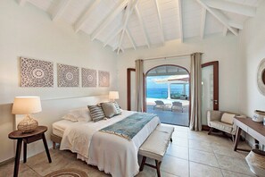 A bedroom with a bed, bedside tables, a bench, and a view of the ocean through a glass door. - Happy Bay   Classic French Creole Villa   Stunning Views (Saint Martin)