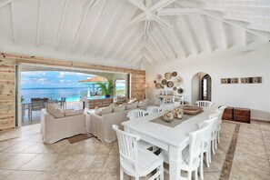 A spacious living area with a large dining table, white chairs, and a view of the ocean through a glass door. - Happy Bay   Classic French Creole Villa   Stunning Views (Saint Martin)