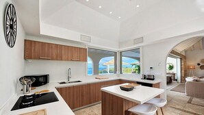 A modern kitchen with wooden cabinets, a white countertop, and a large window offering a view of the outdoors. - Happy Bay   Classic French Creole Villa   Stunning Views (Saint Martin)