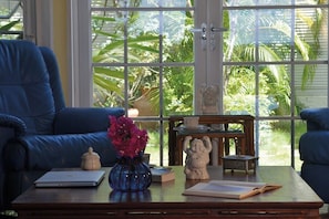 A room with a blue sofa, a wooden coffee table with a laptop, a vase with flowers, and a view of greenery through large windows. - Les Zephyr's   West Indian Style Villa   Stunning Views (Saint Martin)