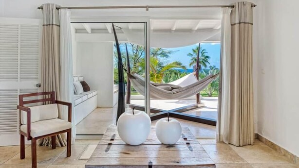 A room with a view of palm trees and the ocean, a hammock, a wooden table with two white vases, and a wooden chair. - La Pinta   Elegant Creole Style Villa (Saint Martin)