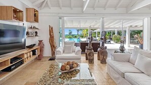 A spacious living room with a flat-screen TV, white sofas, wooden shelves, and a large window overlooking a pool and greenery. - La Pinta   Elegant Creole Style Villa (Saint Martin)