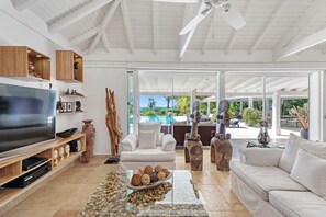 Ein geräumiges Wohnzimmer mit Flachbildfernseher, weißen Sofas, Holzregalen und einem großen Fenster mit Blick auf einen Pool und viel Grün.