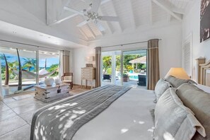Ein geräumiges Schlafzimmer mit einem großen Bett, einem Deckenventilator und Blick auf einen Pool und eine tropische Landschaft durch große Schiebetüren.