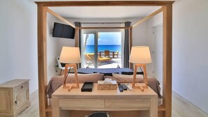 Una habitación con escritorio de madera, dos lámparas, una cama, televisor y vistas a la playa y el océano.