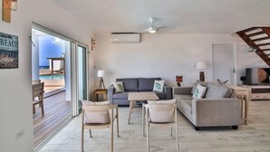 Un salón amplio con sofá, dos sillones, una mesa de centro y televisor. Se observa una escalera que sube a un nivel superior, y un ventanal con vistas a una piscina.