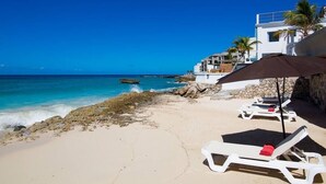 Una playa con sillas de descanso blancas, un muro de piedra y un edificio con balcón con vistas al mar.