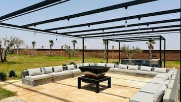 Un patio exterior con un amplio espacio para sentarse, un hogar central y una pérgola en lo alto.
