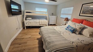 Un dormitorio con una cama litera, una cama individual, un ventilador de techo, un televisor, un aparador y una ventana con persianas.