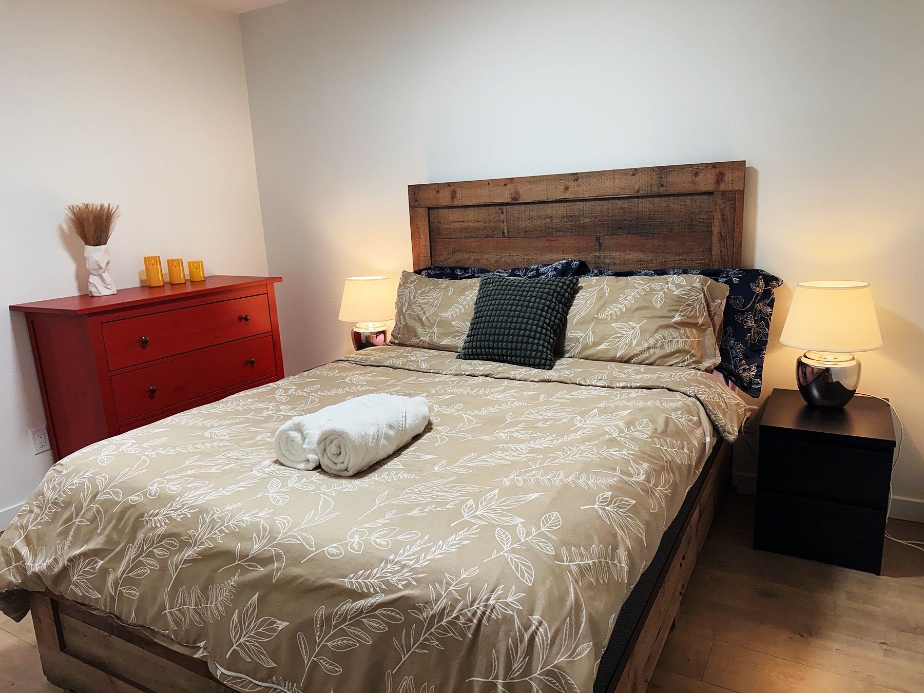 A neatly made bed with a floral bedspread, a wooden headboard, two bedside lamps, a red dresser, and a small vase with dried plants.