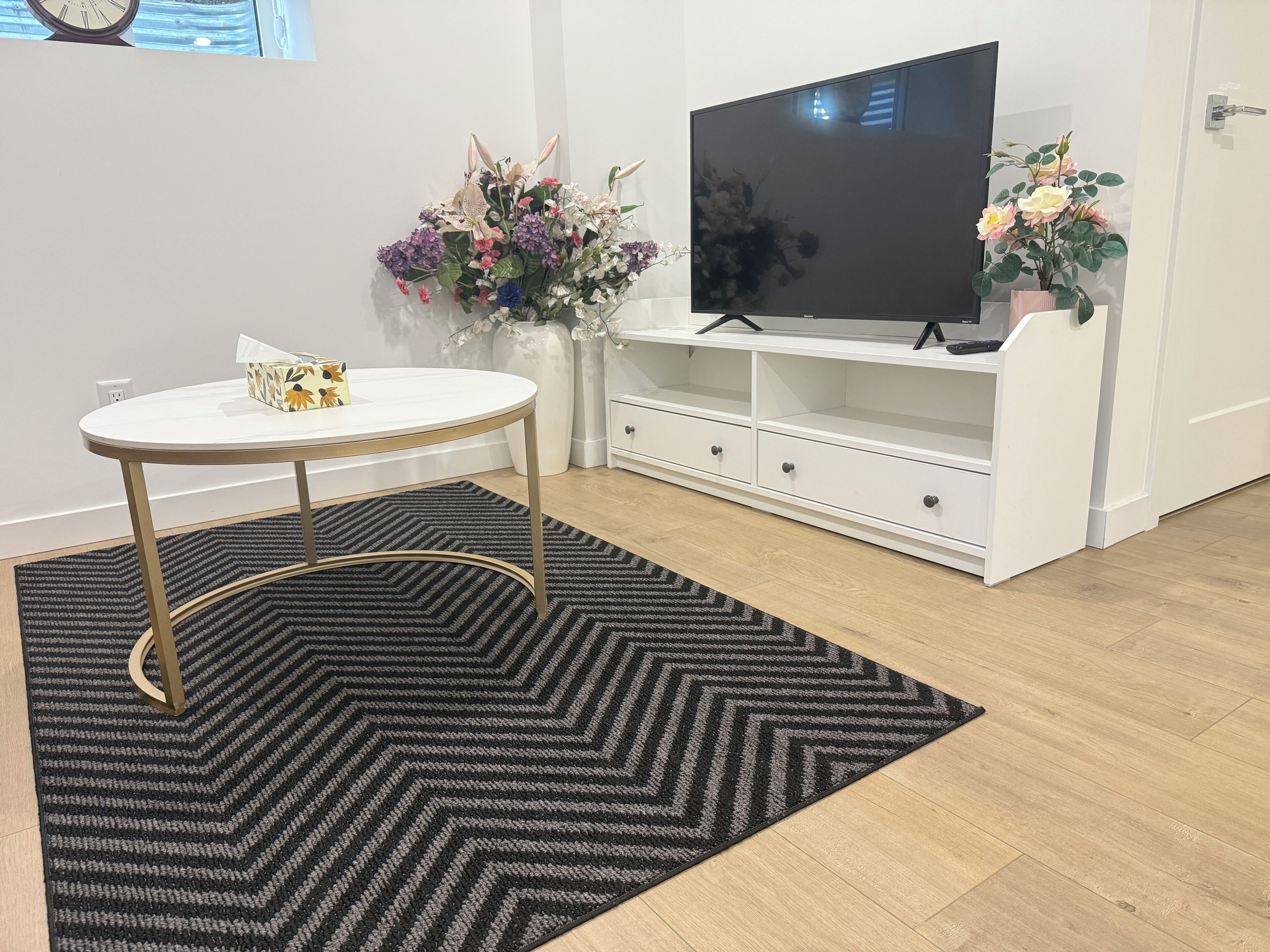 A modern living room with a round white table, a black and gray rug, a white entertainment unit with a flat-screen TV, and two potted plants.