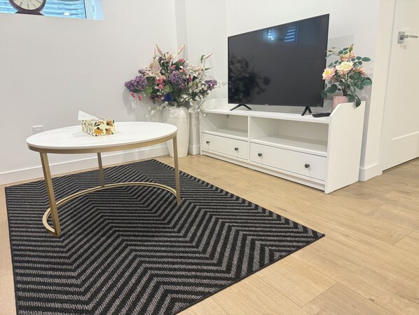 A modern living room with a round white table, a black and gray rug, a white entertainment unit with a flat-screen TV, and two potted plants. - Warm space awaits your experience ! 10minutes walk to reach Langley centre. (Langley Township)