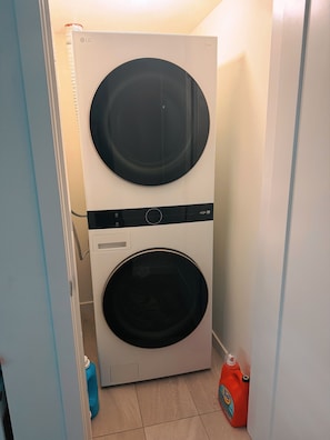 A front-loading white washer and dryer unit with a control panel, located in a narrow hallway with a blue door on the left and a white wall on the right. - Warm space awaits your experience ! 10minutes walk to reach Langley centre. (Langley Township)