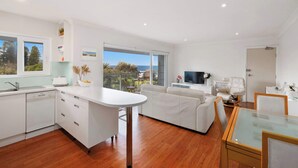 A modern kitchen with a dishwasher, a dining area with a glass table, and a living area with a sofa and TV.