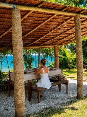 A covered outdoor dining area with a view of the ocean, wooden tables, and a person seated at a table.