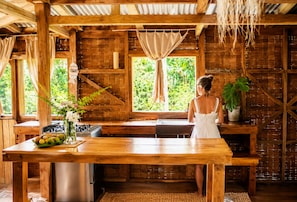 A rustic wooden interior with a large wooden table, a kitchen area, and a view of greenery outside.