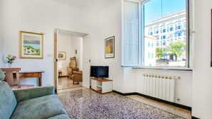 Una stanza con una grande finestra, una televisione, un divano, un piccolo tavolino con un vaso e un portafoto appeso al muro.