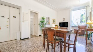 Una camera d'albergo con un tavolo da pranzo in legno, sedie, una televisione e un'ampia finestra che si affaccia su edifici.