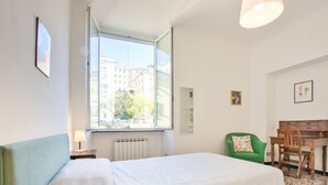 Una camera da letto con un letto, una finestra vista edifici, una sedia verde, una scrivania in legno e un termosifone.