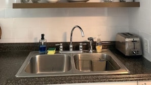 Eine Küche mit doppelter Spüle, einem Edelstahlarmaturen und einem Toaster. Darüber befinden sich zwei Holzregale, auf denen sich Glaswaren und Geschirr sowie ein Topf mit Pflanze befinden.