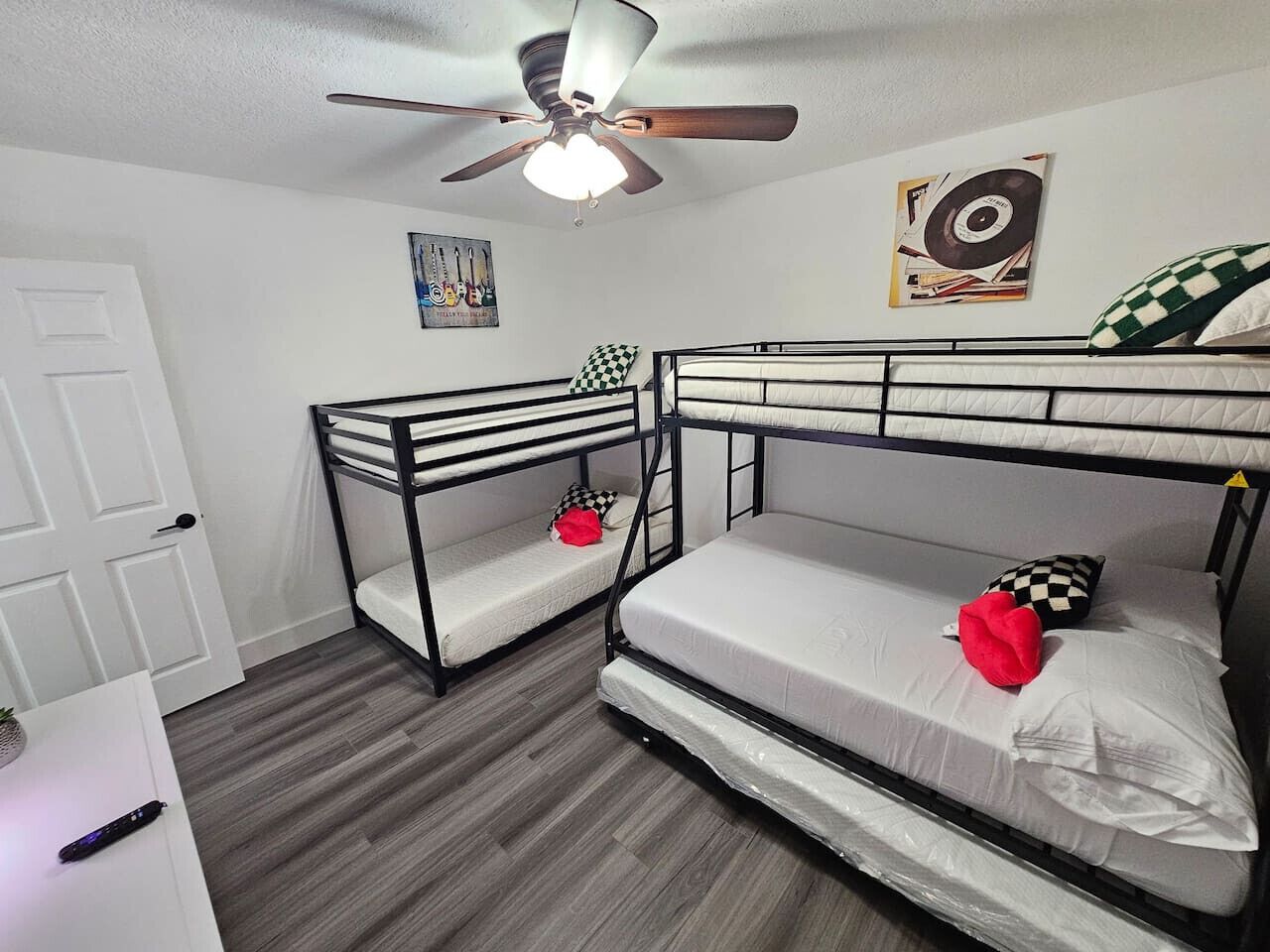 A room with bunk beds, a ceiling fan, a door, and a small table with a remote control.