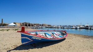 A beach with a rowboat on the sand, a clear blue sky, and a marina in the background. - Stunning apartment in Marzamemi (Marzamemi)
