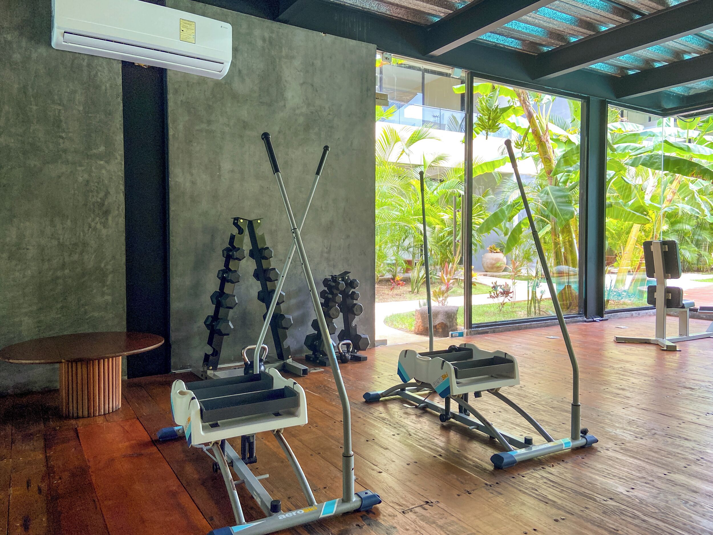 A gym with cardio equipment, a round wooden table, and a view of a tropical garden through large windows.