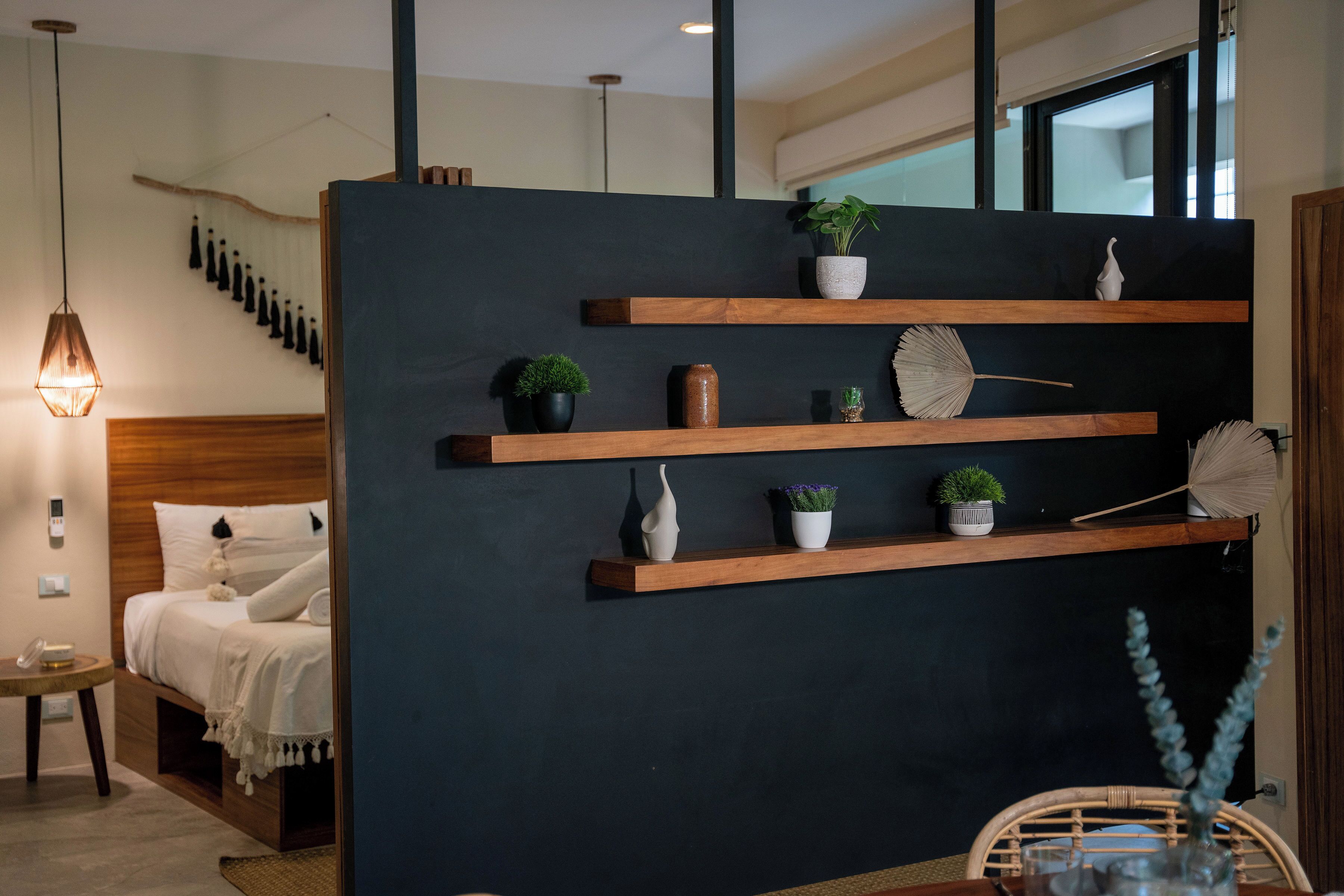 Une chambre d’hôtel moderne avec un mur d’accent de couleur sombre, des étagères en bois ornées d’objets décoratifs, un lit avec une literie blanche, une table de chevet avec une lampe, et une vue sur l’extérieur par une fenêtre.