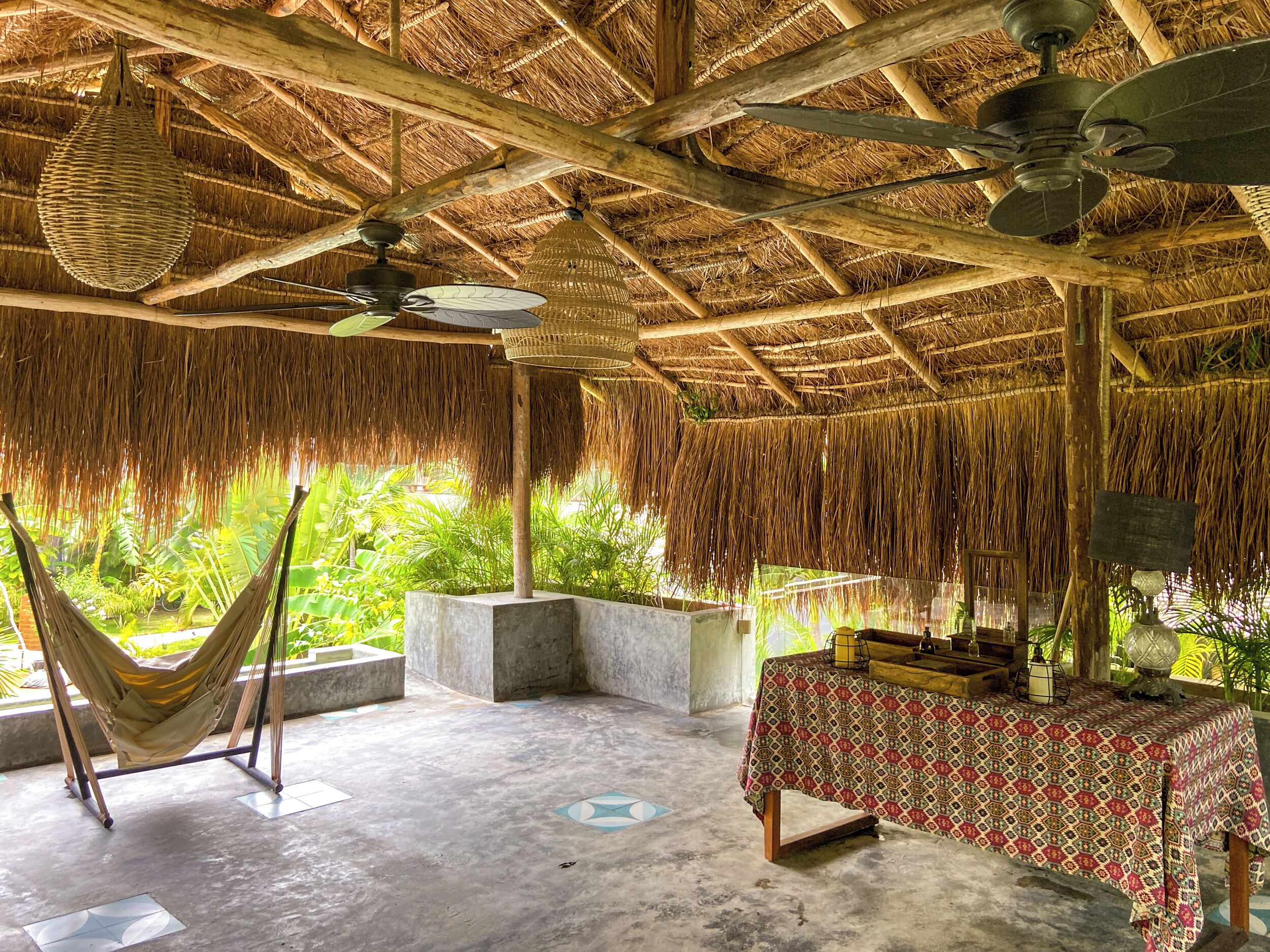 A thatched-roof room with a hammock, a table with a patterned tablecloth, and a ceiling fan.